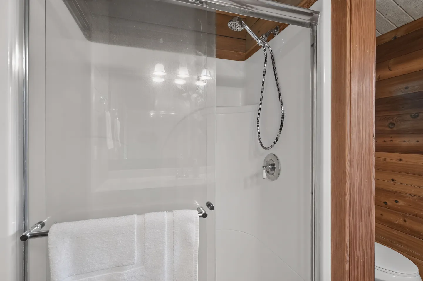 A close-up view of a bathroom shower stall with a glass door, chrome handheld showerhead, and white towel hanging on the handle.