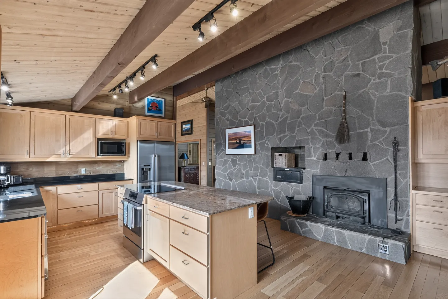 Kitchen with light wood cabinetry, an island, and a tall stone fireplace wall with a wood-burning insert.