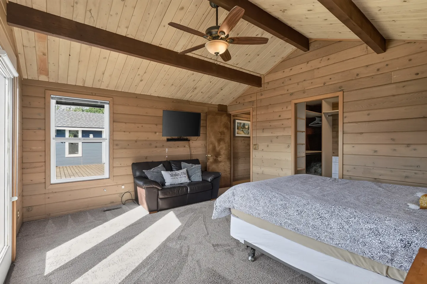 Bedroom with wood-paneled walls and ceiling, a bed, a small leather sofa beneath a wall-mounted TV, and a window.
