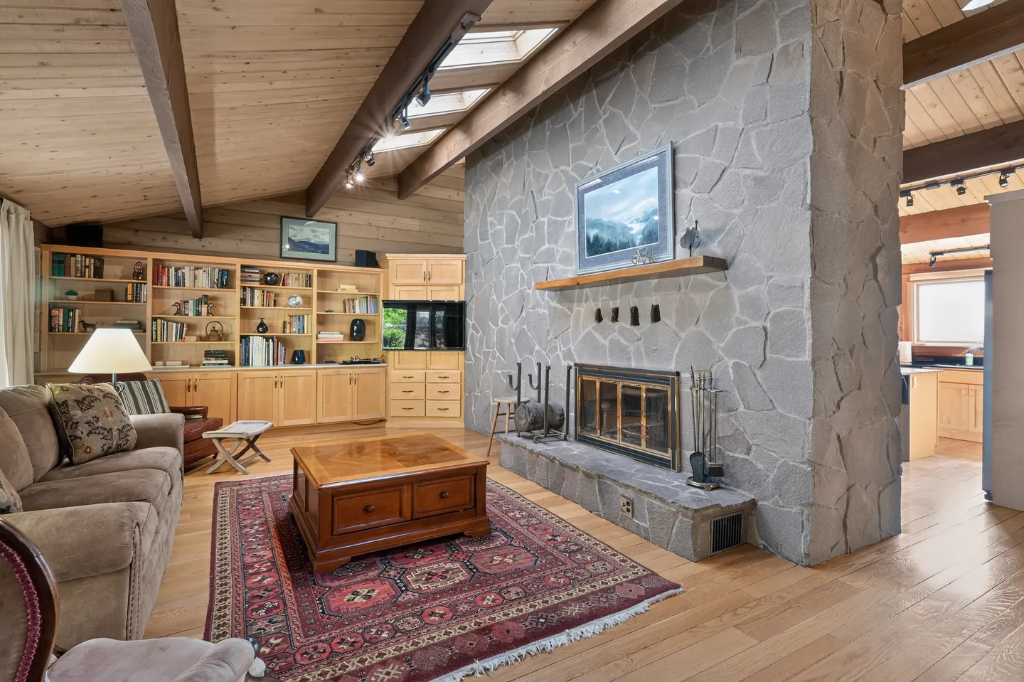 Living room with a large stone fireplace, wood-paneled vaulted ceiling with skylights, built-in bookcases, and comfortable seating.