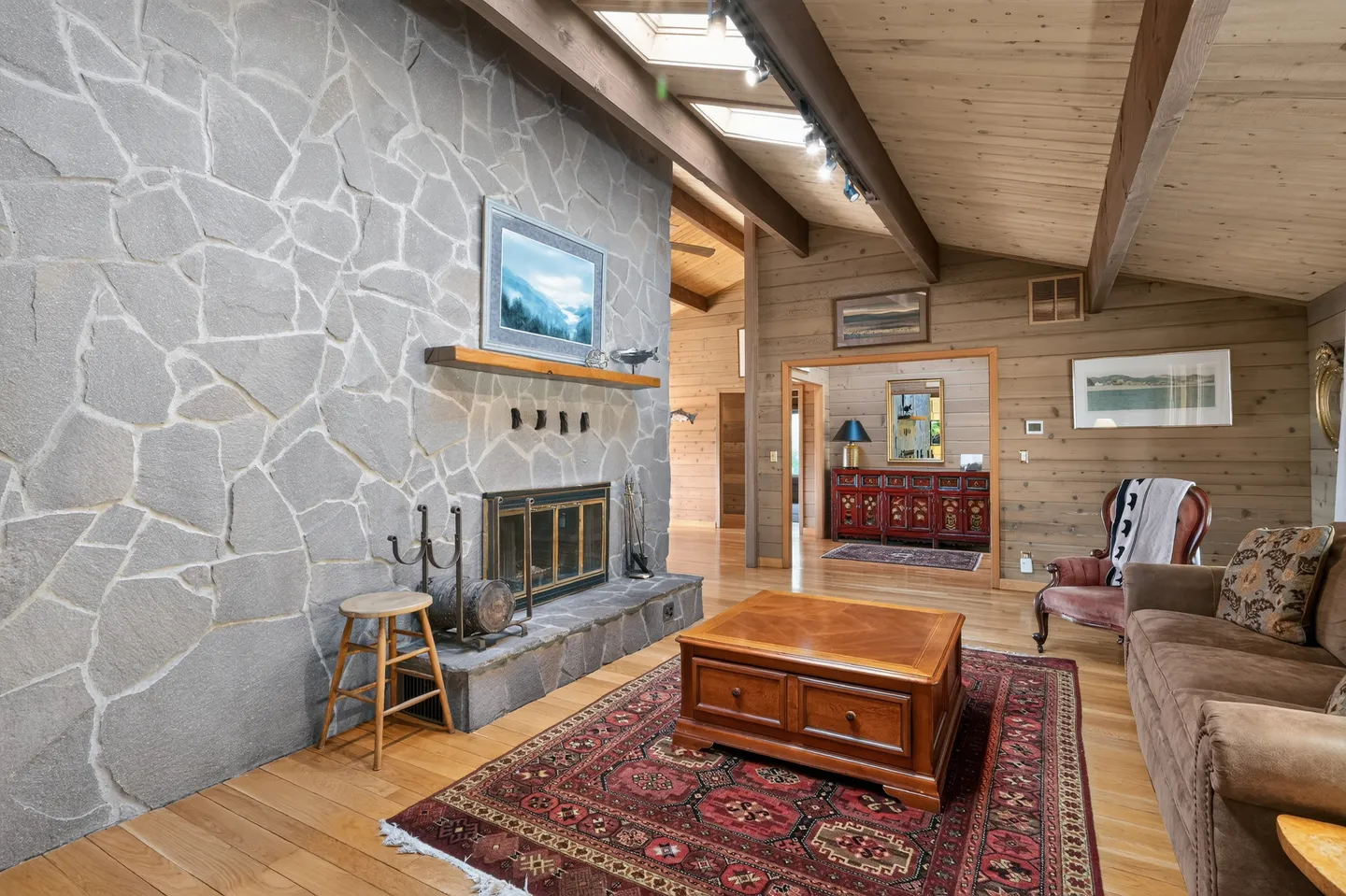 Living room with a large stone fireplace, wood-paneled walls, vaulted ceiling with exposed beams, and a patterned rug under a wooden coffee table.