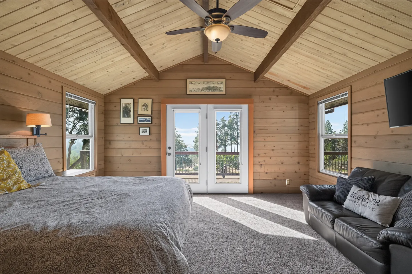 Bedroom with wood-paneled walls, vaulted ceiling with exposed beams, carpet, bed, sofa, and glass French doors leading to a balcony.
