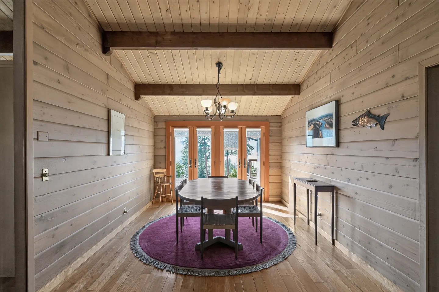 Dining room with wood paneled walls, wood ceiling with exposed beams, oval dining table, purple rug, and glass double doors at the far end.