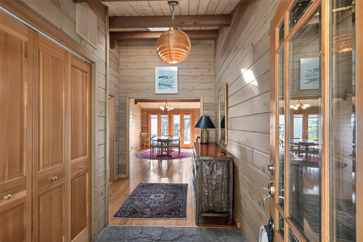 Entryway with wood-paneled walls and ceiling, a round hanging pendant lamp, and a view into the dining area.