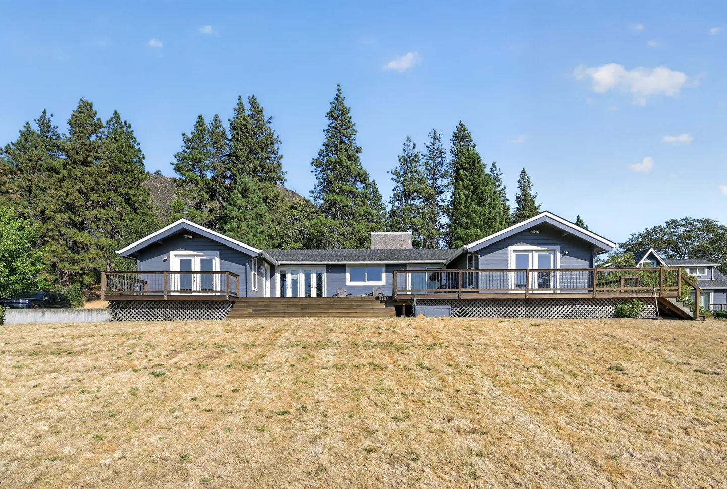 Exterior rear view of a one-story blue house with an expansive wooden deck facing a dry, grassy yard backed by tall pine trees.