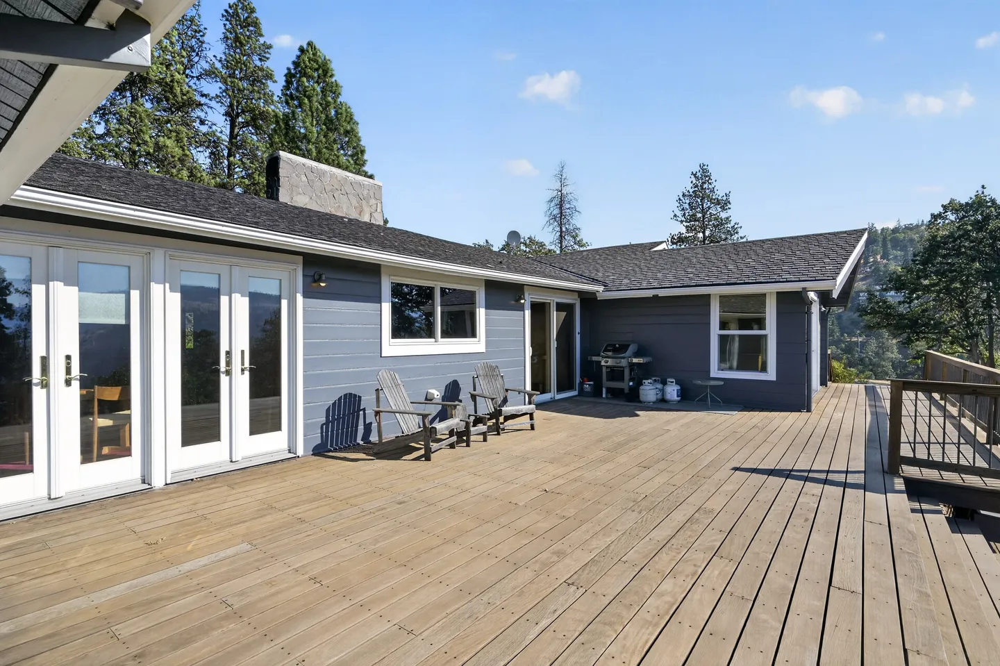 Spacious outdoor wooden deck with patio chairs, barbecue grill, and views of surrounding trees and blue sky.