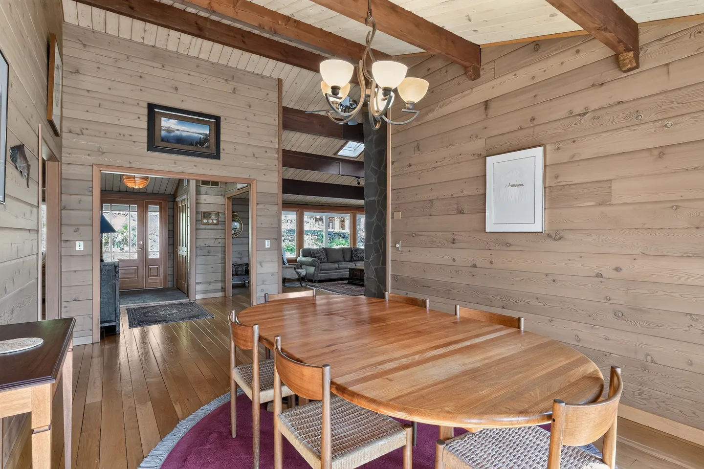 Dining area with a large oval wooden table and chairs, wood-paneled walls, exposed ceiling beams, and an opening showing the foyer and living room.