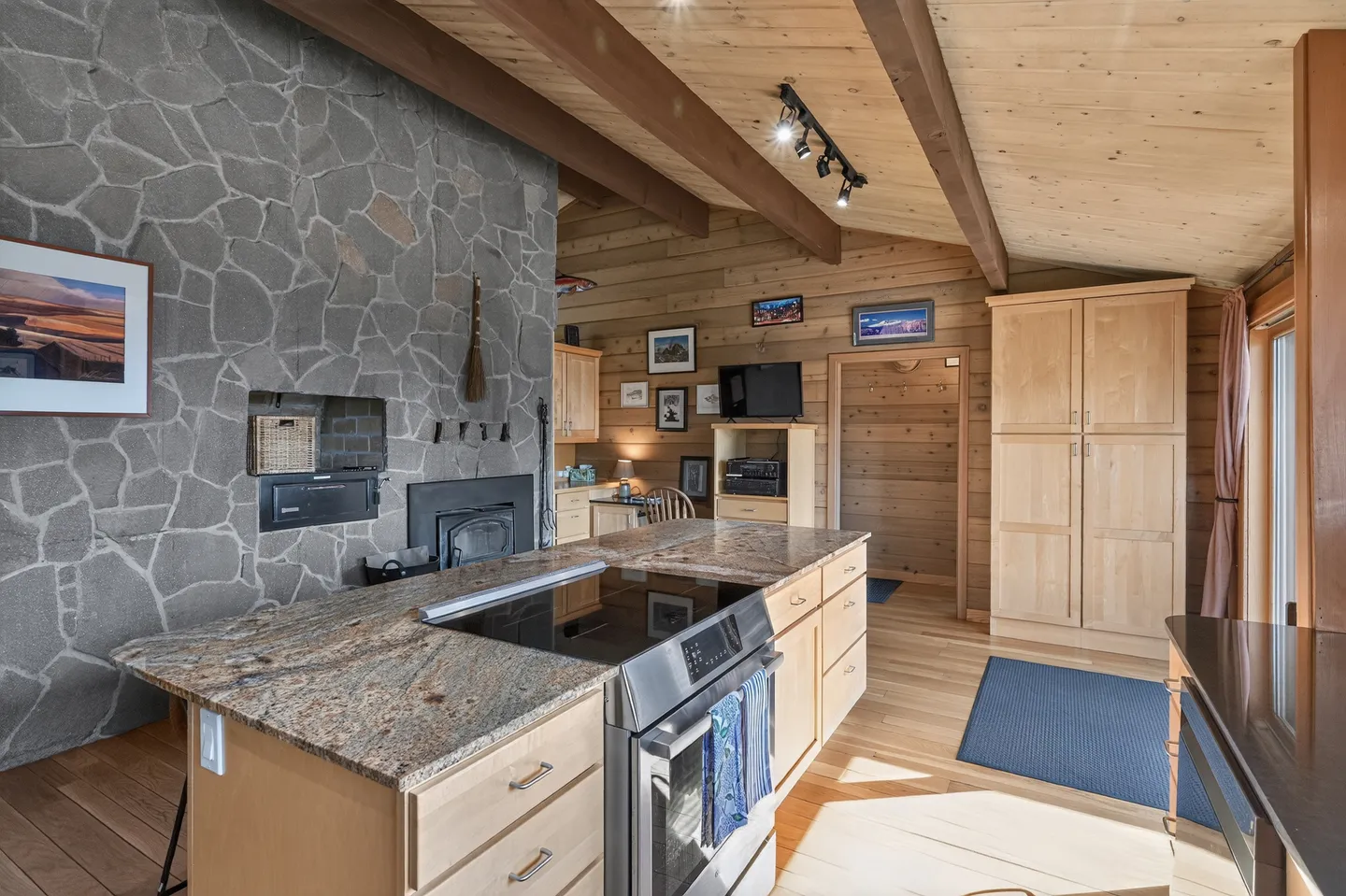 Kitchen island with electric stove top and granite counter adjacent to a tall stone fireplace in a wood-paneled room.
