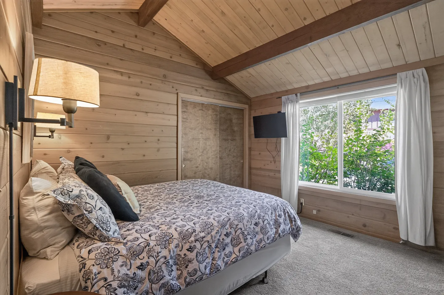 Bedroom with wood-paneled walls and ceiling, a bed with patterned bedding, a wall-mounted TV, and a window framed by white curtains.