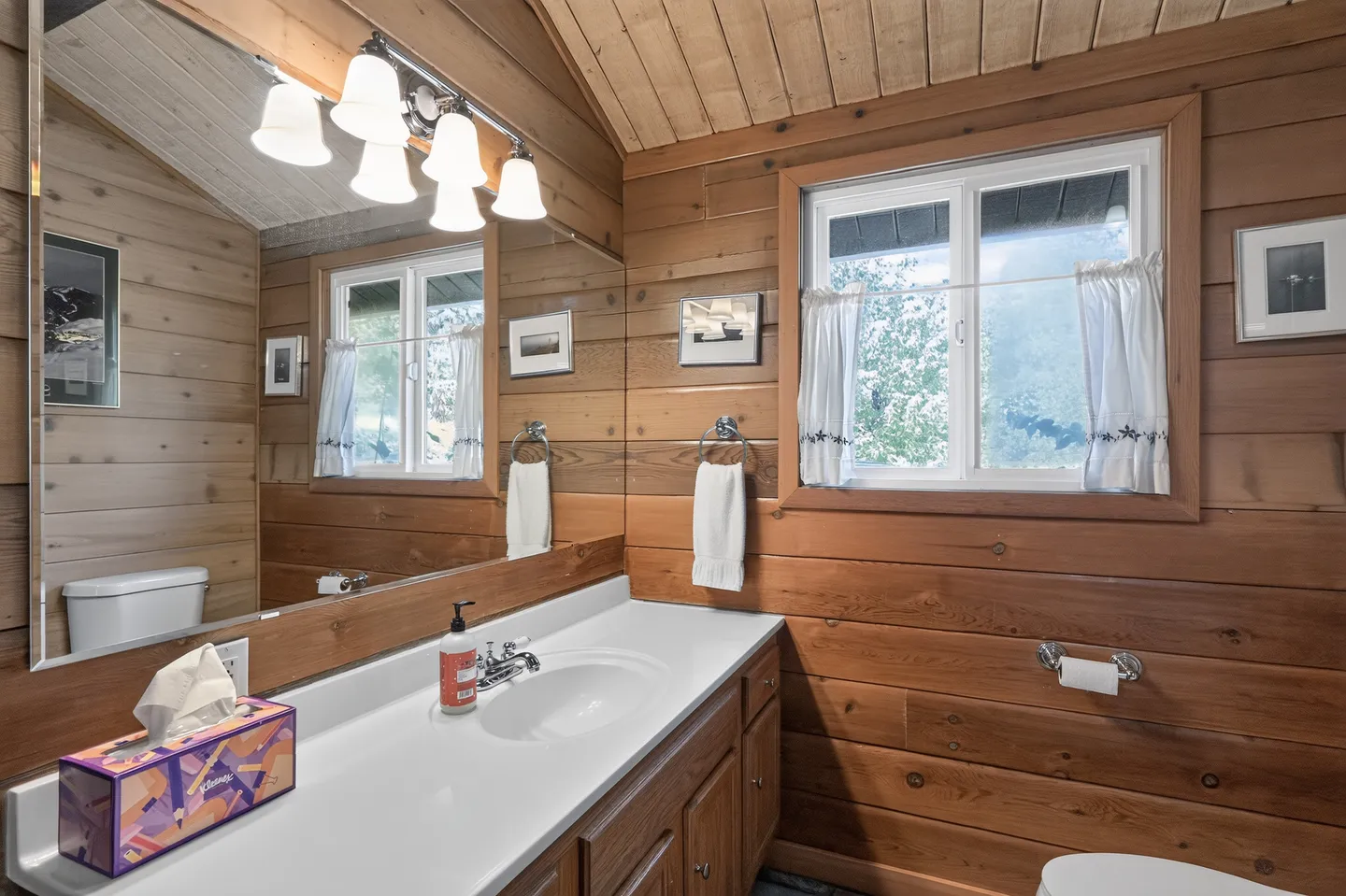 Bathroom with wood-paneled walls, vanity with white countertop, large mirror, and a window with small white curtains.