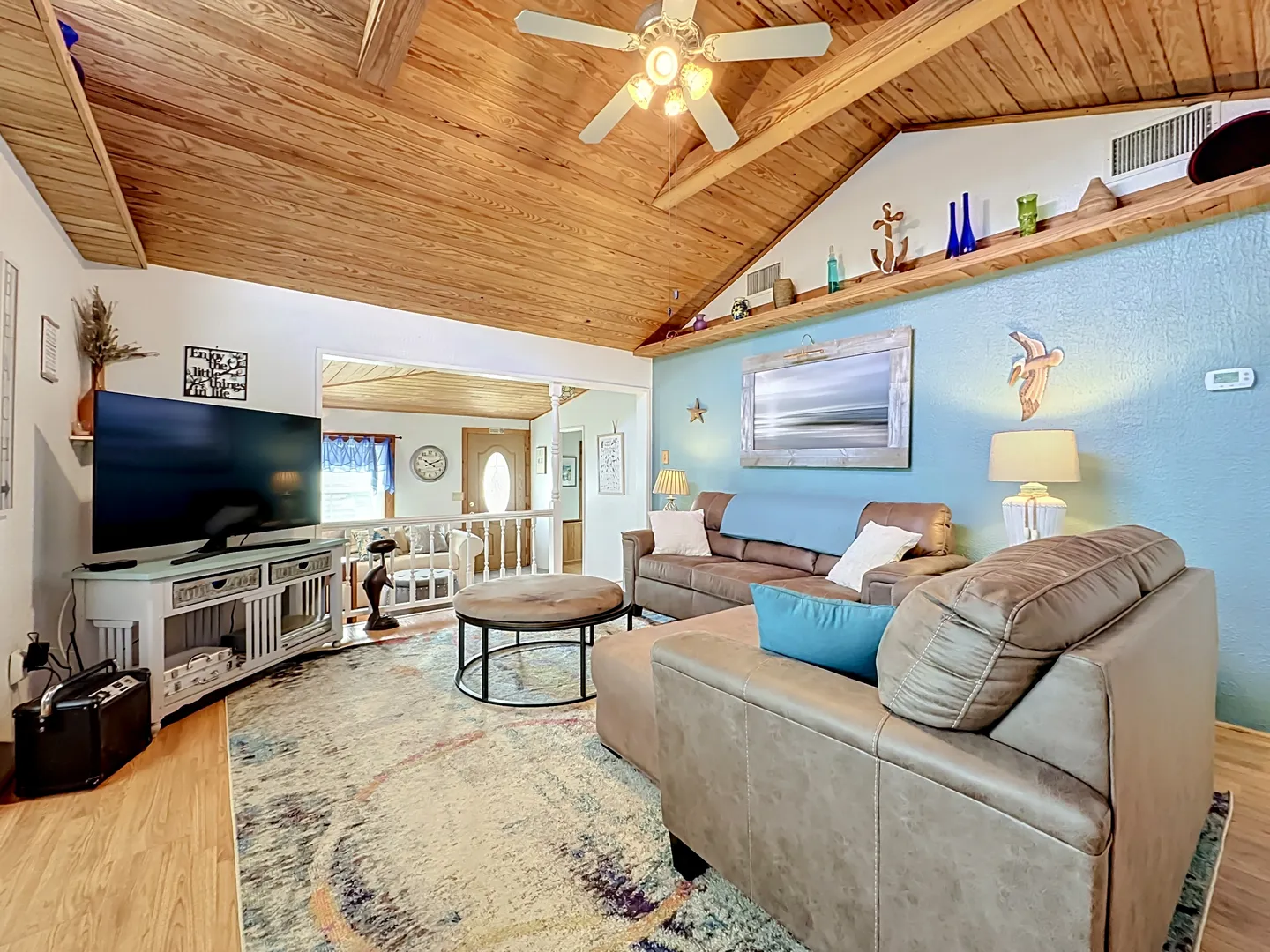 Living room featuring a vaulted wood-paneled ceiling with exposed beams, ceiling fan, brown sectional sofa, TV on a stand, and blue accent wall.