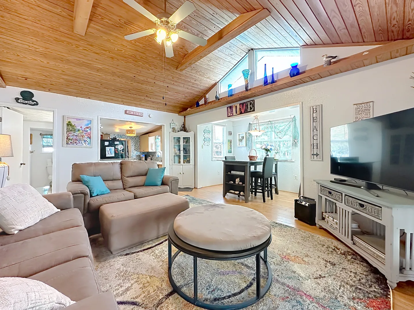 Open-concept living room with wood-paneled vaulted ceiling, ceiling fan, sofa seating, and television, opening into the dining area and kitchen.