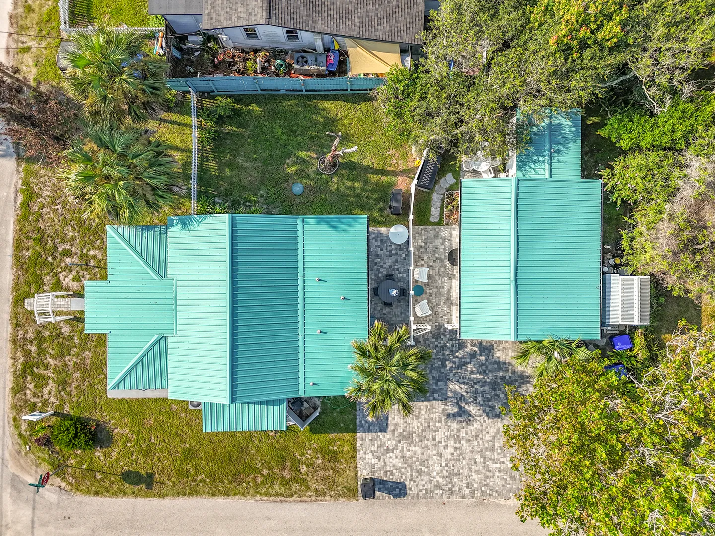 Aerial overhead view showing the property with teal metal roofs, a paved driveway and patio, outdoor seating, and lush tropical trees.