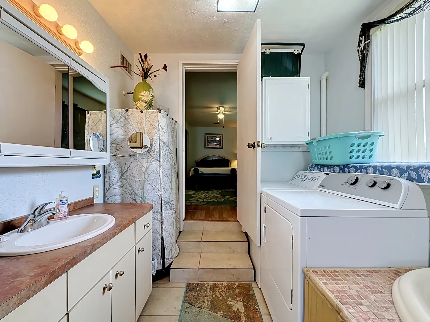 Bathroom and laundry area with vanity, washing machine and dryer, leading up steps to a bedroom.