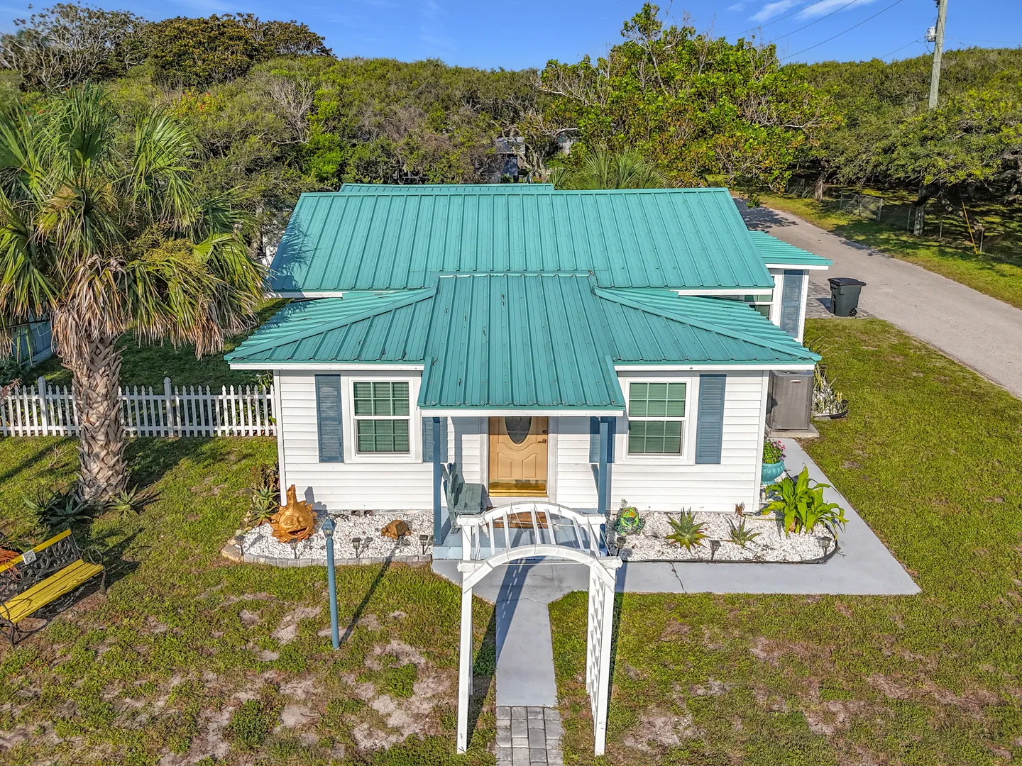 Elevated view of a white cottage with a teal metal roof, white entry arbor, and palm tree.