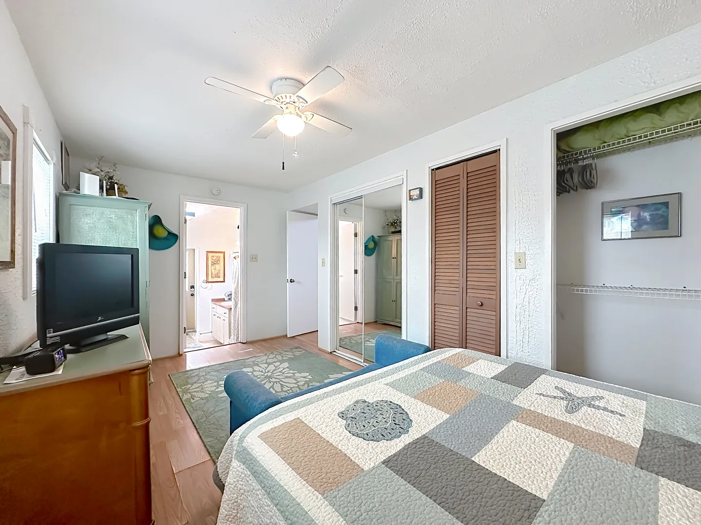 Bedroom with patterned quilt bed, television on a dresser, ceiling fan, mirrored closet doors, and open door leading to a bathroom.