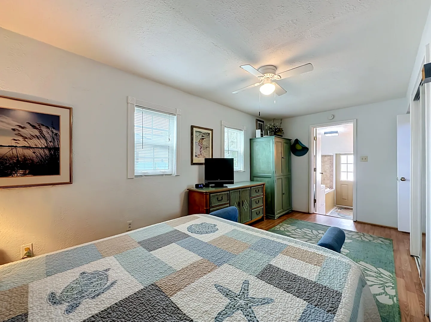 Bedroom with coastal-themed quilt, dresser with television, tall green cabinet, and view into an ensuite bathroom.