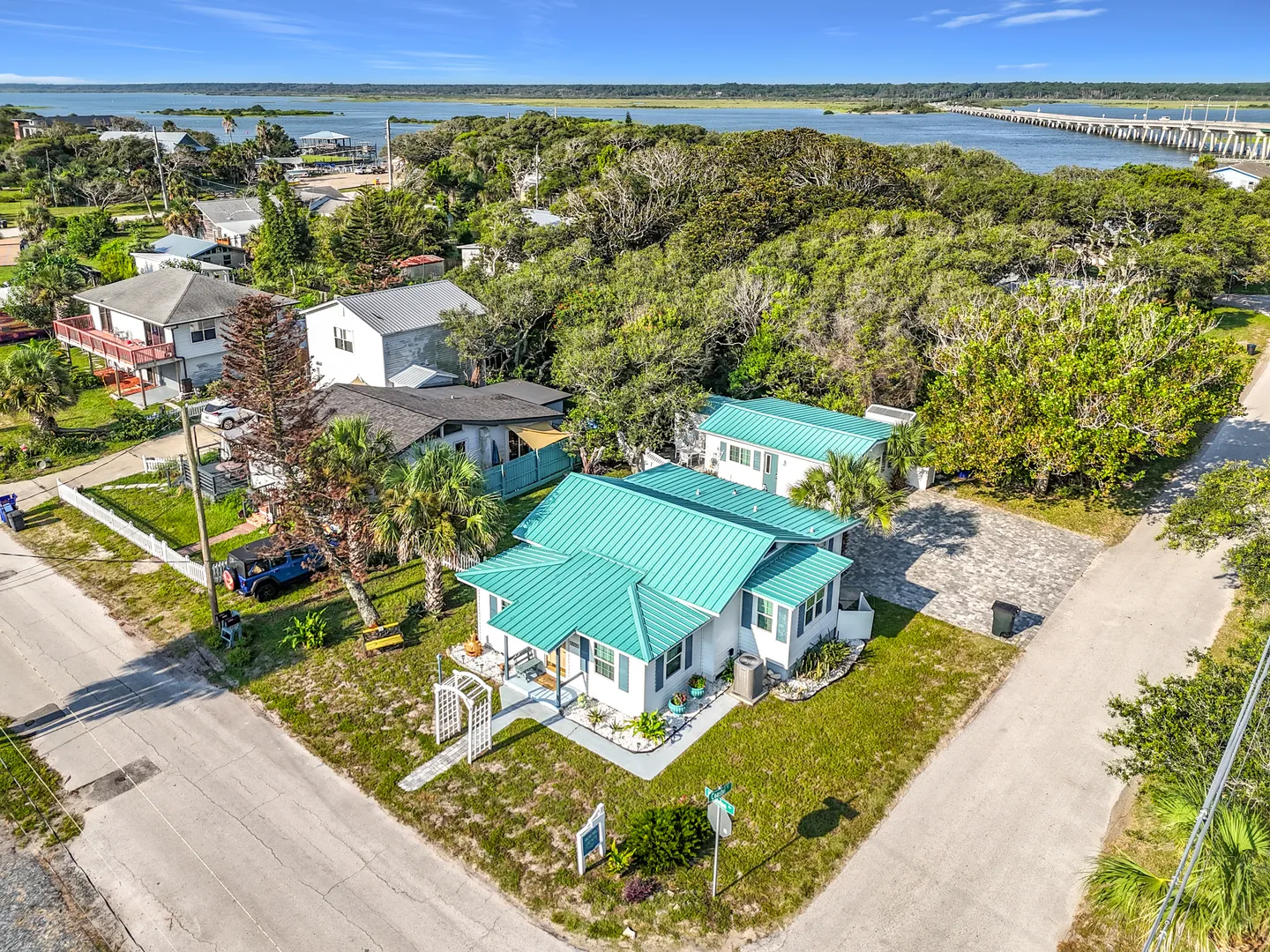 Aerial view of a coastal cottage with a bright teal metal roof near lush trees, roads, and a large body of water with a bridge in the distance.