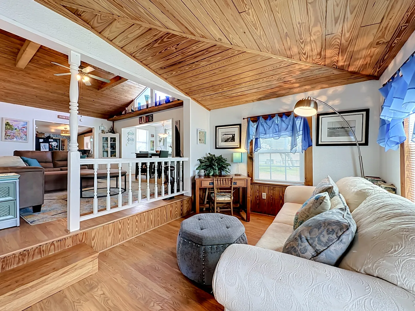 Sunken sitting room with wood-paneled ceiling, cream sofa, and steps leading up to an adjacent living area behind a white railing.