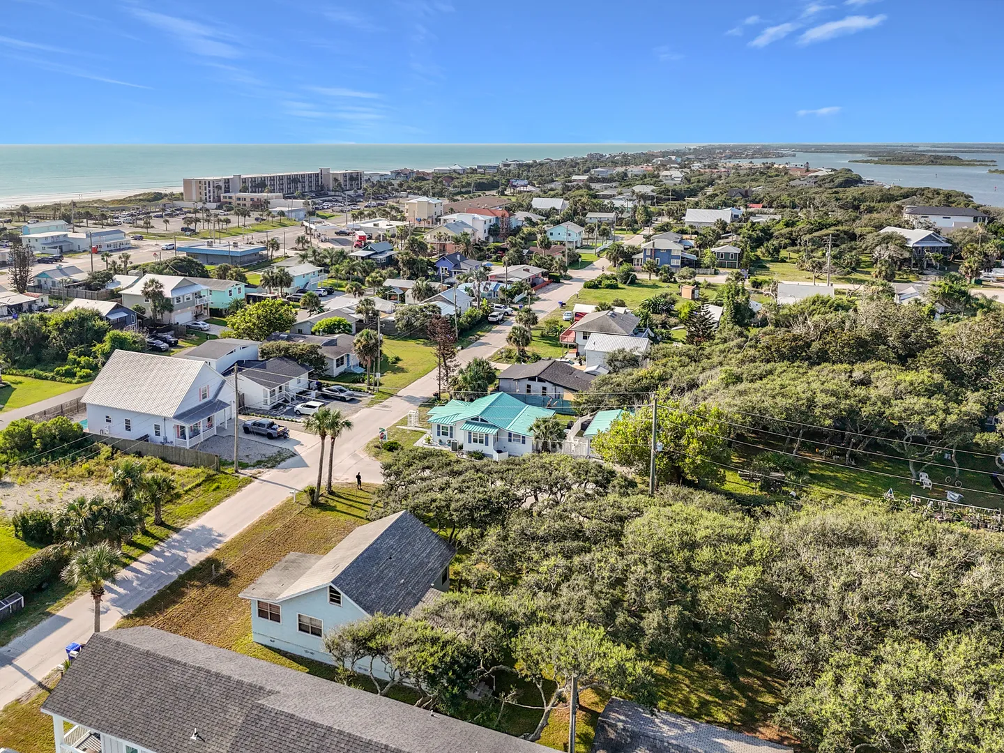 Aerial view of a coastal neighborhood with houses, palm trees, and the ocean visible on the horizon under a clear blue sky.