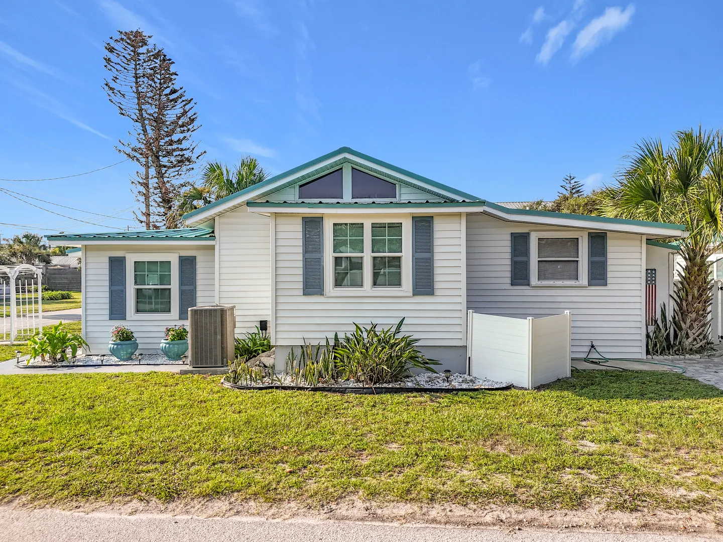 Exterior front view of a single-story white home with blue shutters, a green roof trim, and a green lawn under a blue sky.