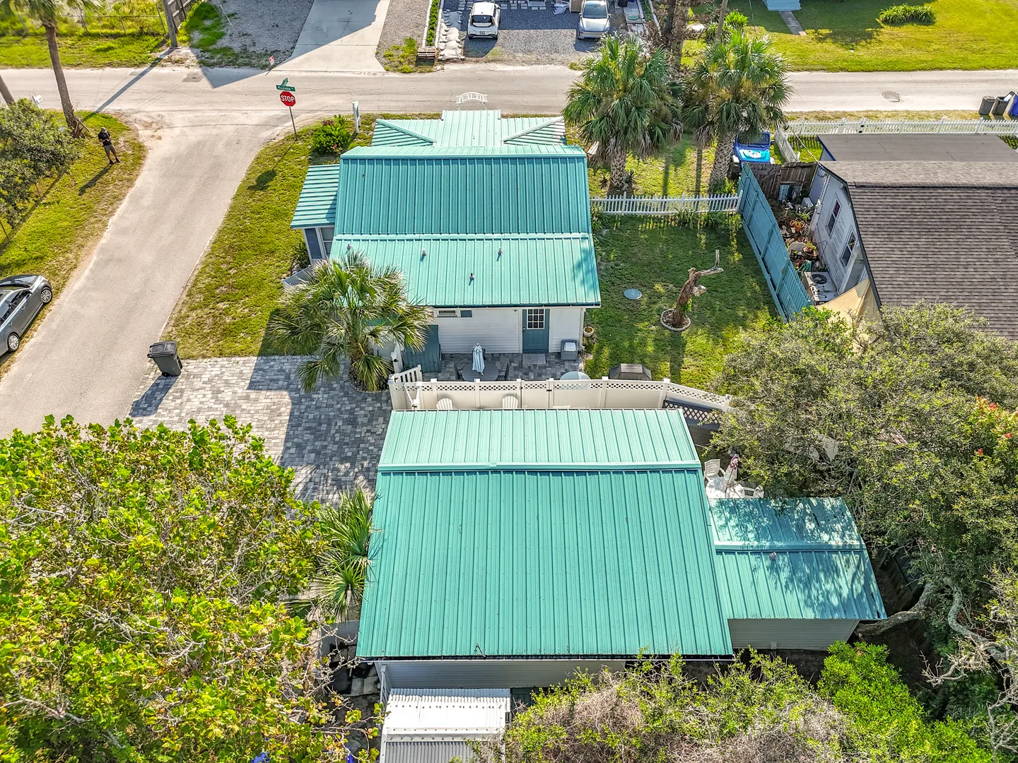 Aerial view of a coastal cottage property with bright green metal roofs, paved parking, and a fenced yard with palm trees.