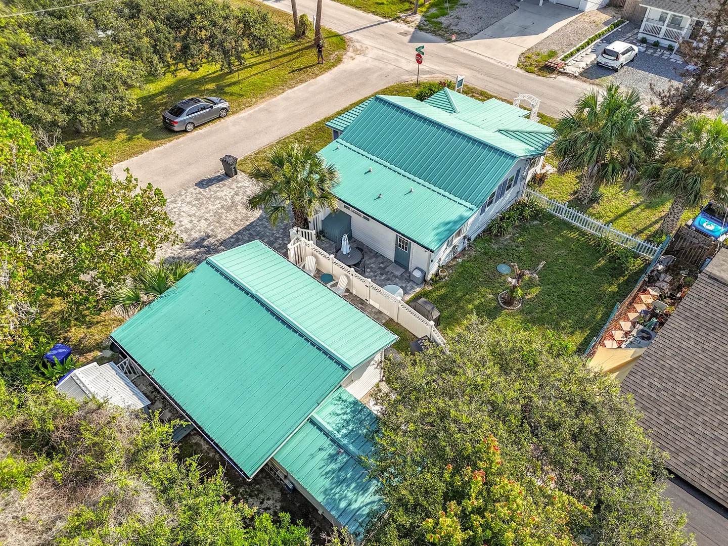 Aerial view of a coastal cottage property with bright turquoise metal roofs, a fenced yard, and surrounding palm trees.