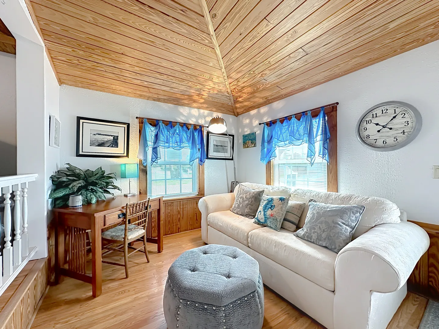Living area featuring a white sofa with accent pillows, a round tufted ottoman, a wooden desk with a chair, and a vaulted wood-paneled ceiling.