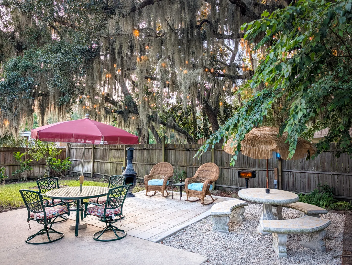 Outdoor patio with multiple seating areas, umbrellas, and rocking chairs beneath trees draped in Spanish moss and string lights.