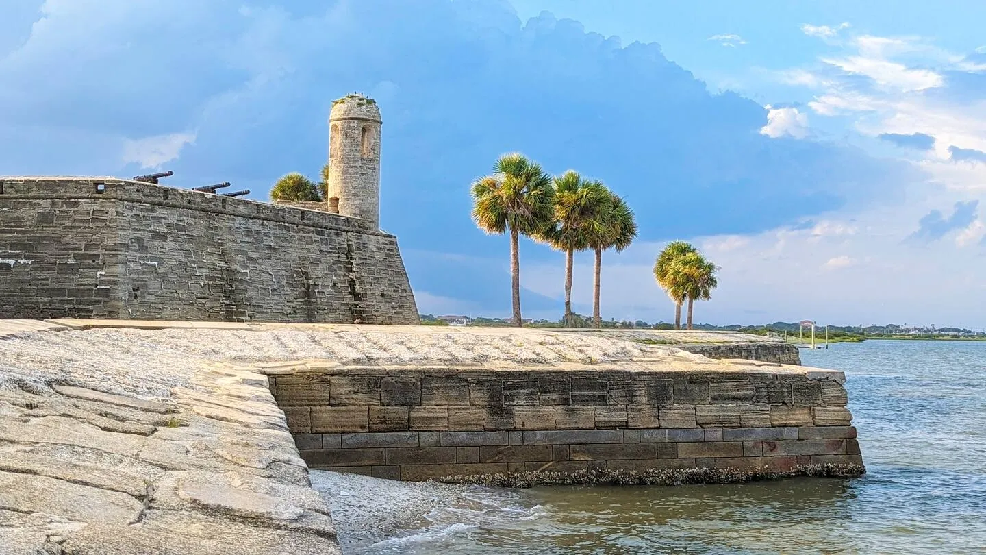 A historic stone masonry fort with a watchtower and cannons along a waterfront under a partly cloudy sky with palm trees.
