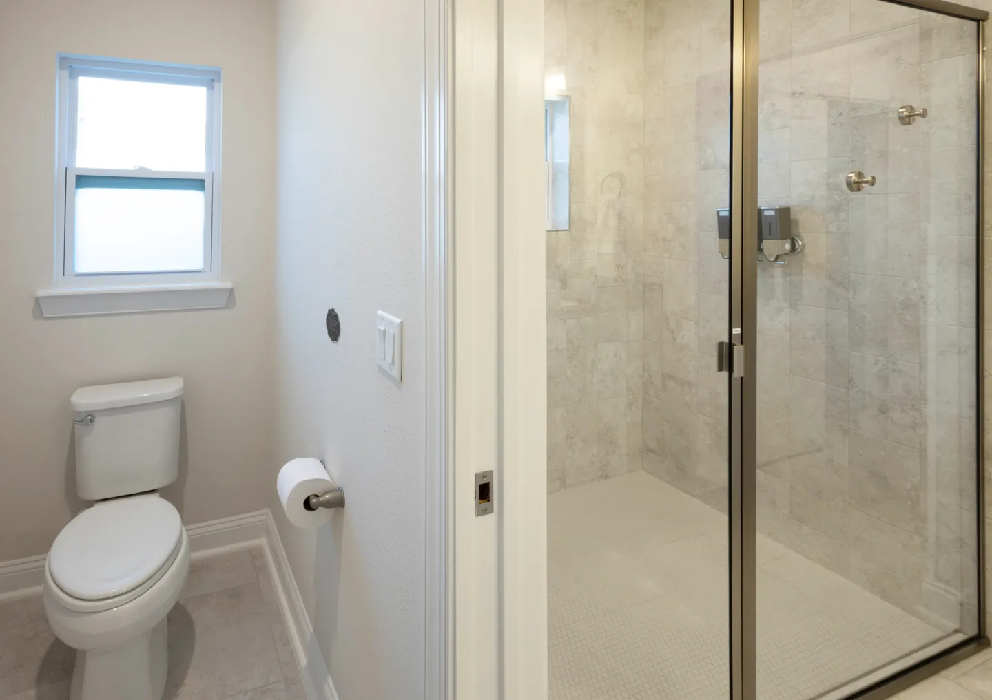 Bathroom showing a white toilet near a frosted window and an adjacent walk-in shower with glass doors and tiled walls.