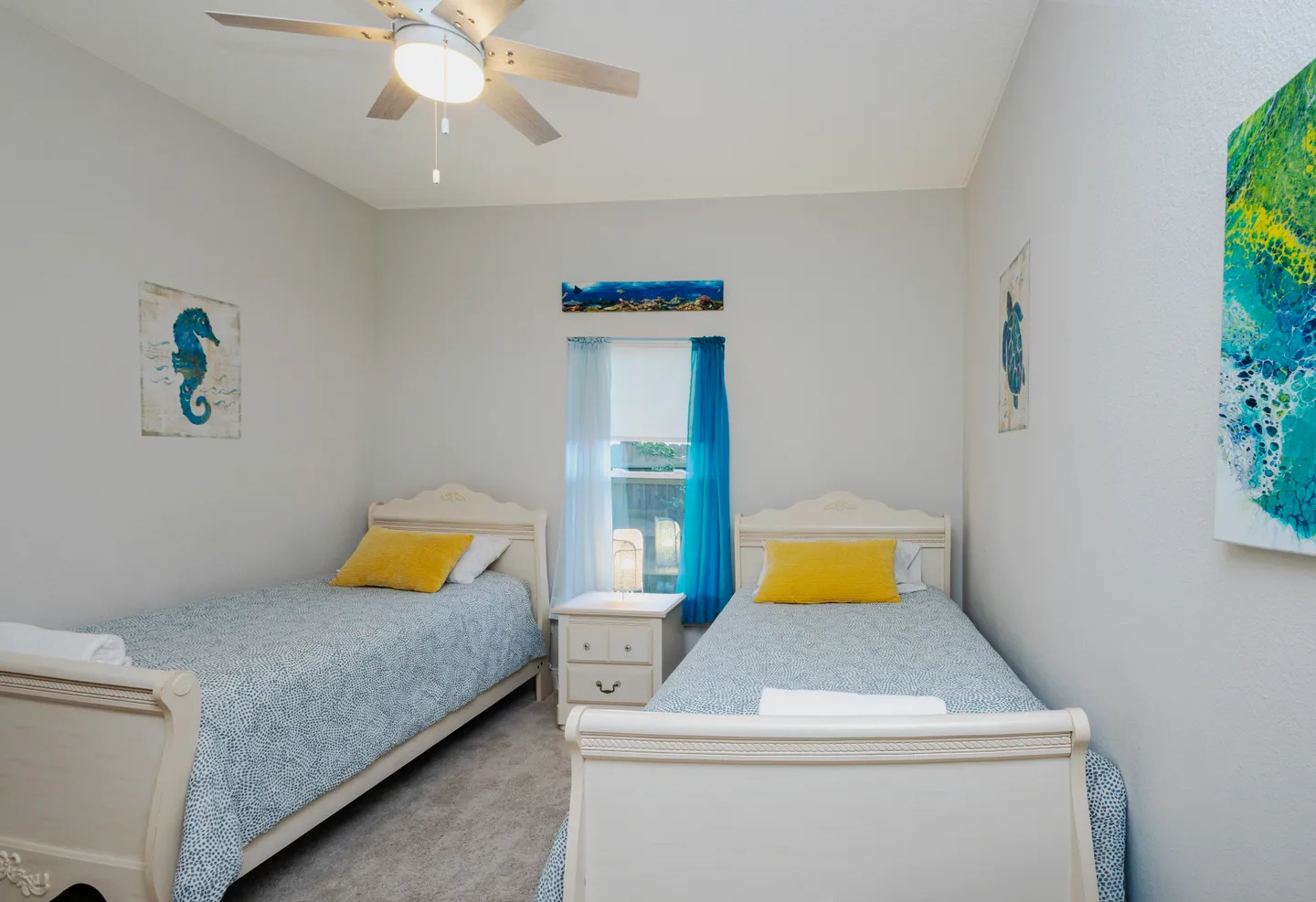 Bedroom featuring two twin beds with white frames, patterned blue bedding, yellow accent pillows, a central nightstand, and ocean-themed wall art.
