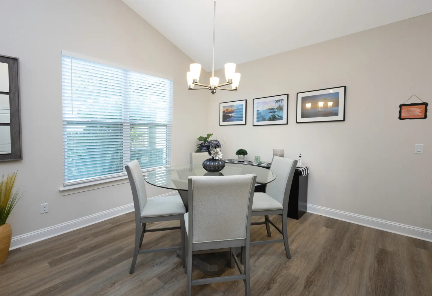 Dining area with a round glass table, four upholstered chairs, and a chandelier below a vaulted ceiling.