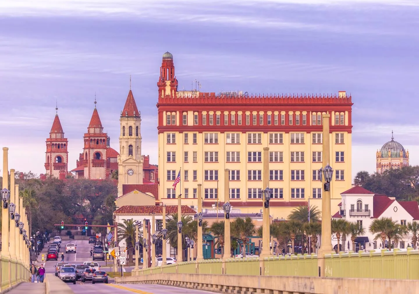 Elevated view across a bridge with lamp posts leading toward historic city architecture and towers under a pale sky.