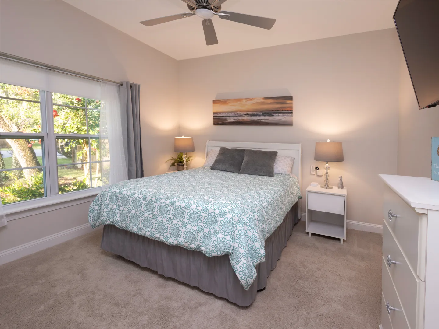 Bedroom with a bed dressed in a patterned coverlet, carpeted floor, ceiling fan, and window looking outside.
