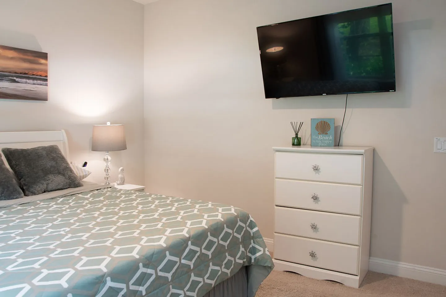 Bedroom featuring a bed with patterned bedding, a bedside table with a lamp, a white four-drawer dresser, and a wall-mounted flat-screen television.