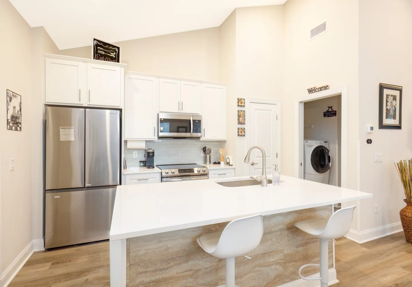 Kitchen with white cabinetry, stainless steel appliances, and a breakfast bar island with two modern stools, opening to a laundry room.