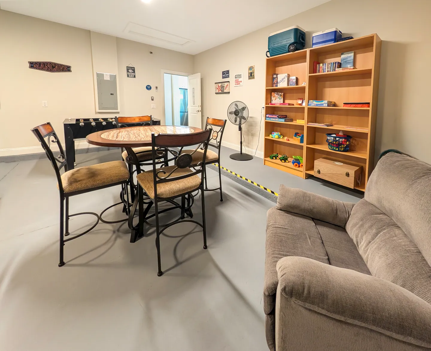 Game room setup in a garage space featuring a round table with four chairs, a foosball table, an armchair, and wooden bookcases stocked with board games and toys.