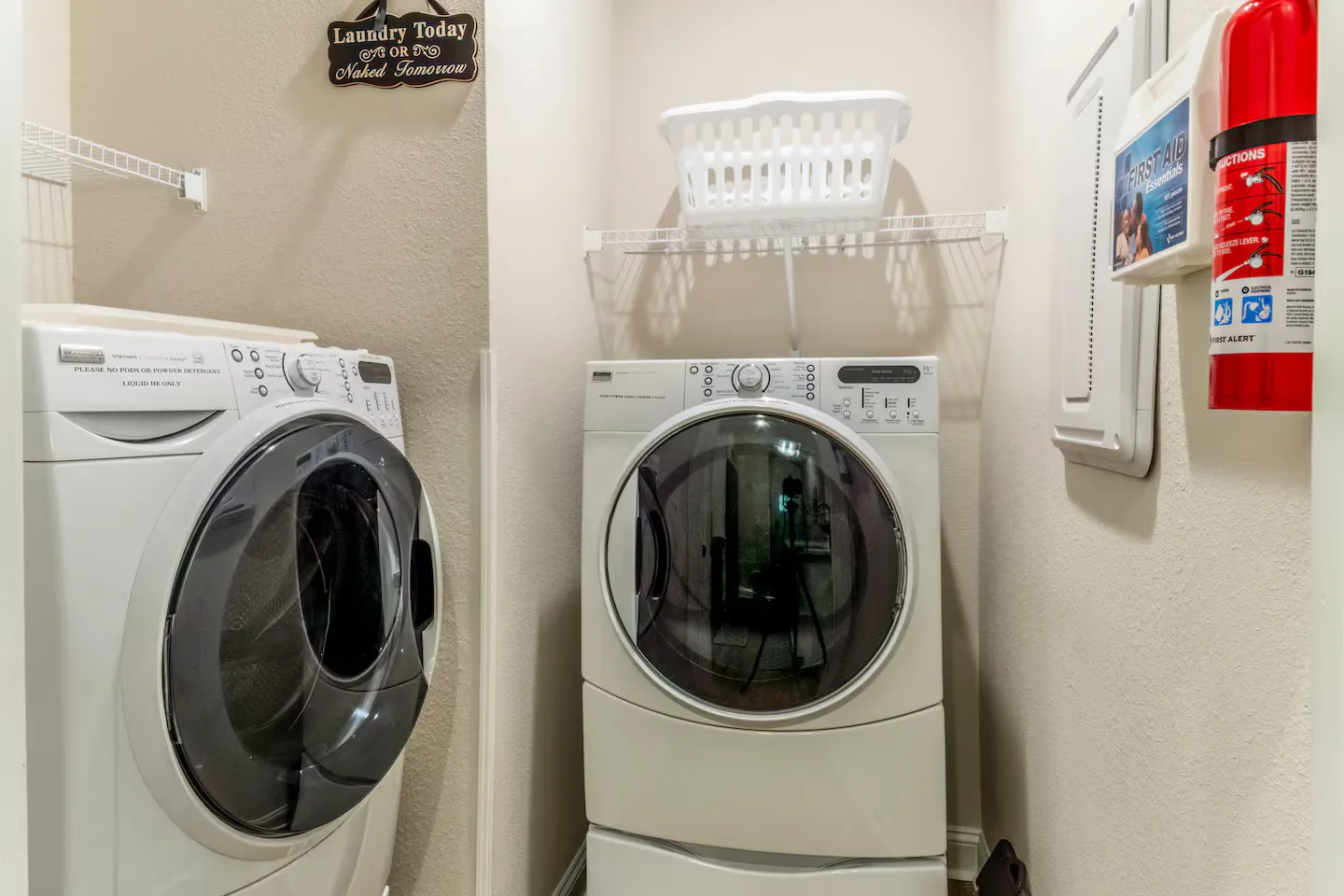 A laundry alcove featuring a white front-loading washer and dryer, wire shelving, a laundry basket, and safety equipment.