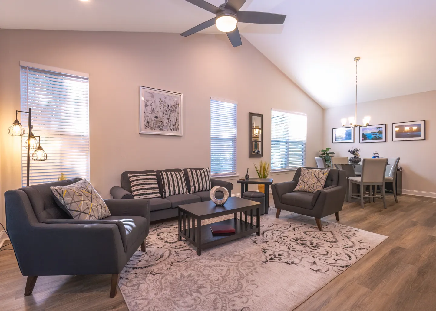 Living room with high vaulted ceiling, dark grey sofa and armchairs arranged around a patterned area rug and coffee table, adjacent to a dining area.