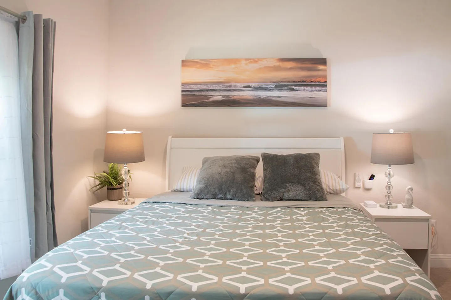 Bedroom with a patterned bedspread, white headboard, matching nightstands with lamps, and a beach panoramic artwork on the wall.