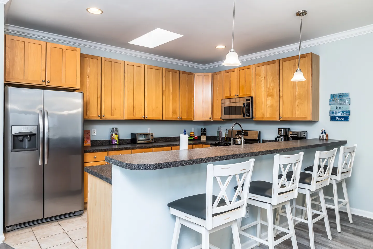 Kitchen with light wood cabinets, dark countertops, a raised breakfast bar with four white barstools, and stainless steel appliances.