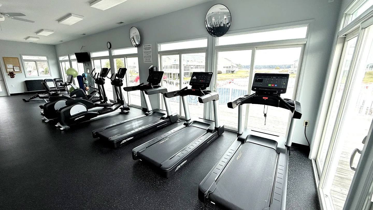 An indoor fitness room featuring treadmills and elliptical machines lined up facing large sliding glass windows.