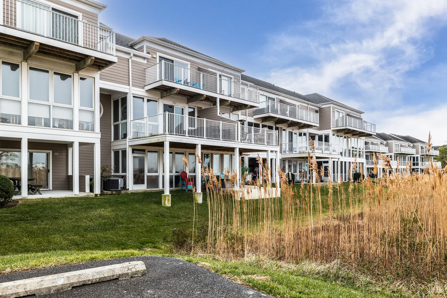 Exterior view of multi-level townhomes or condominiums with white balconies and decks overlooking a grassy lawn with tall reeds.