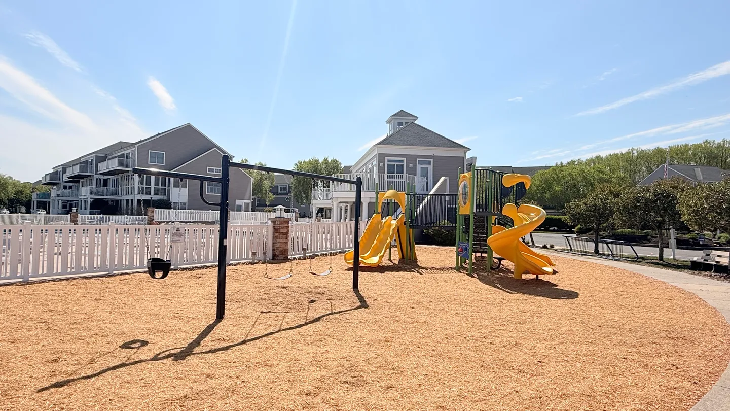 Outdoor community playground with yellow slides and swing sets surrounded by wood mulch on a sunny day.