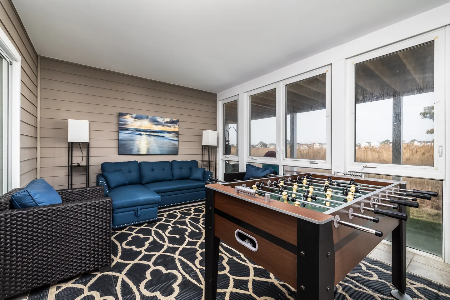 Sunroom with a foosball table, blue sectional sofa, wicker armchair, patterned rug, and large windows.