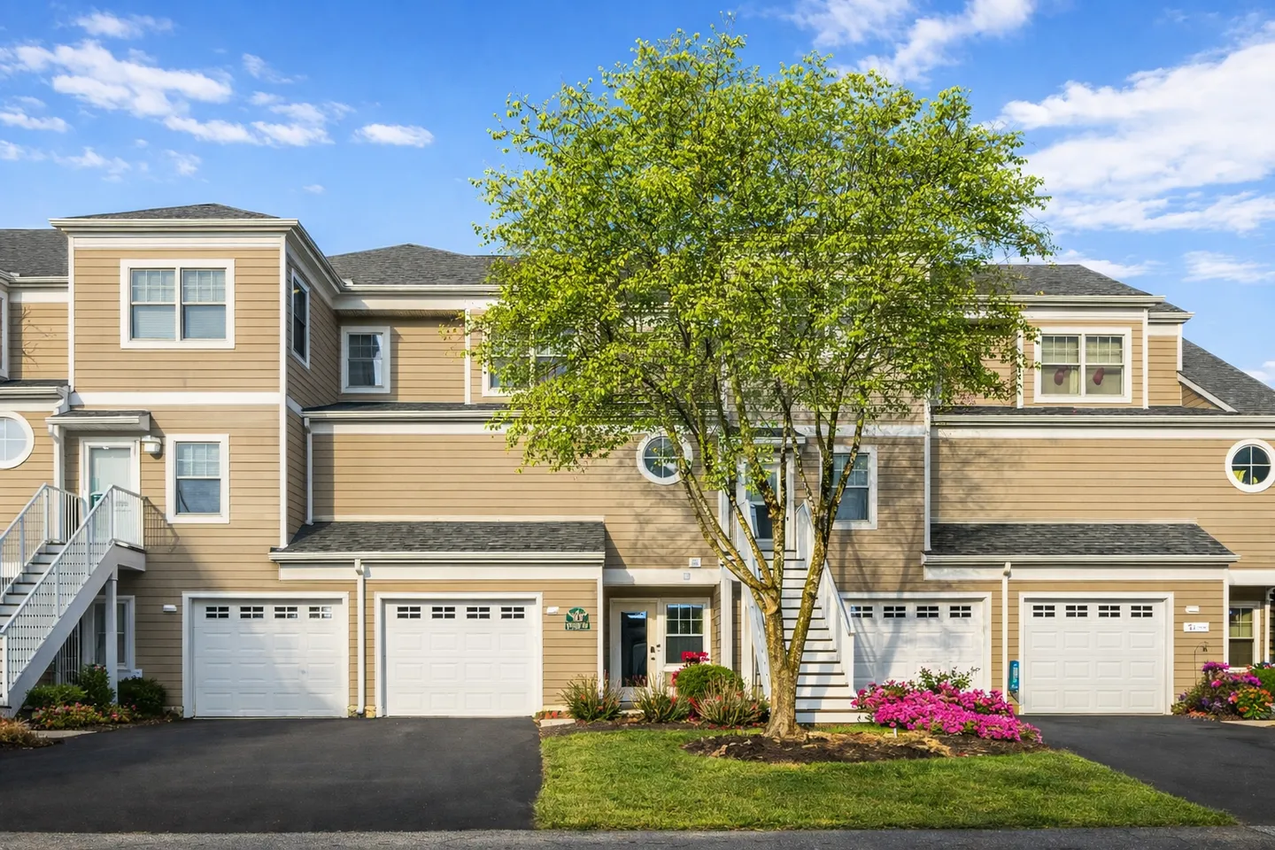 Exterior view of multi-level beige townhomes with white garages, exterior stairs, and a leafy green tree in front.