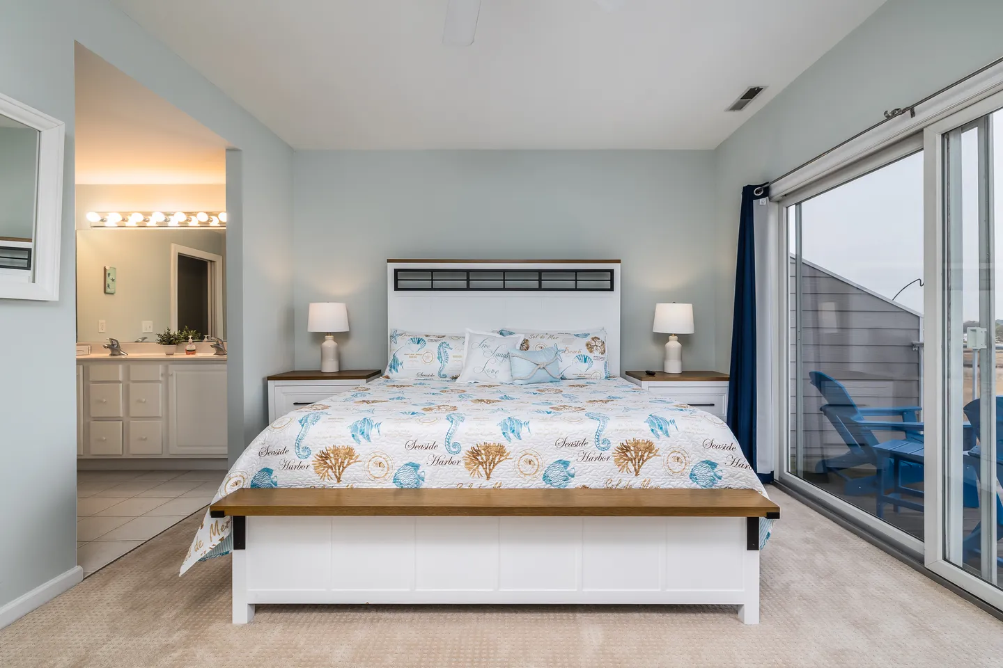 Bedroom with coastal-themed bedding, a white and wood bed frame, sliding glass doors to a balcony, and an en-suite bathroom visible to the left.