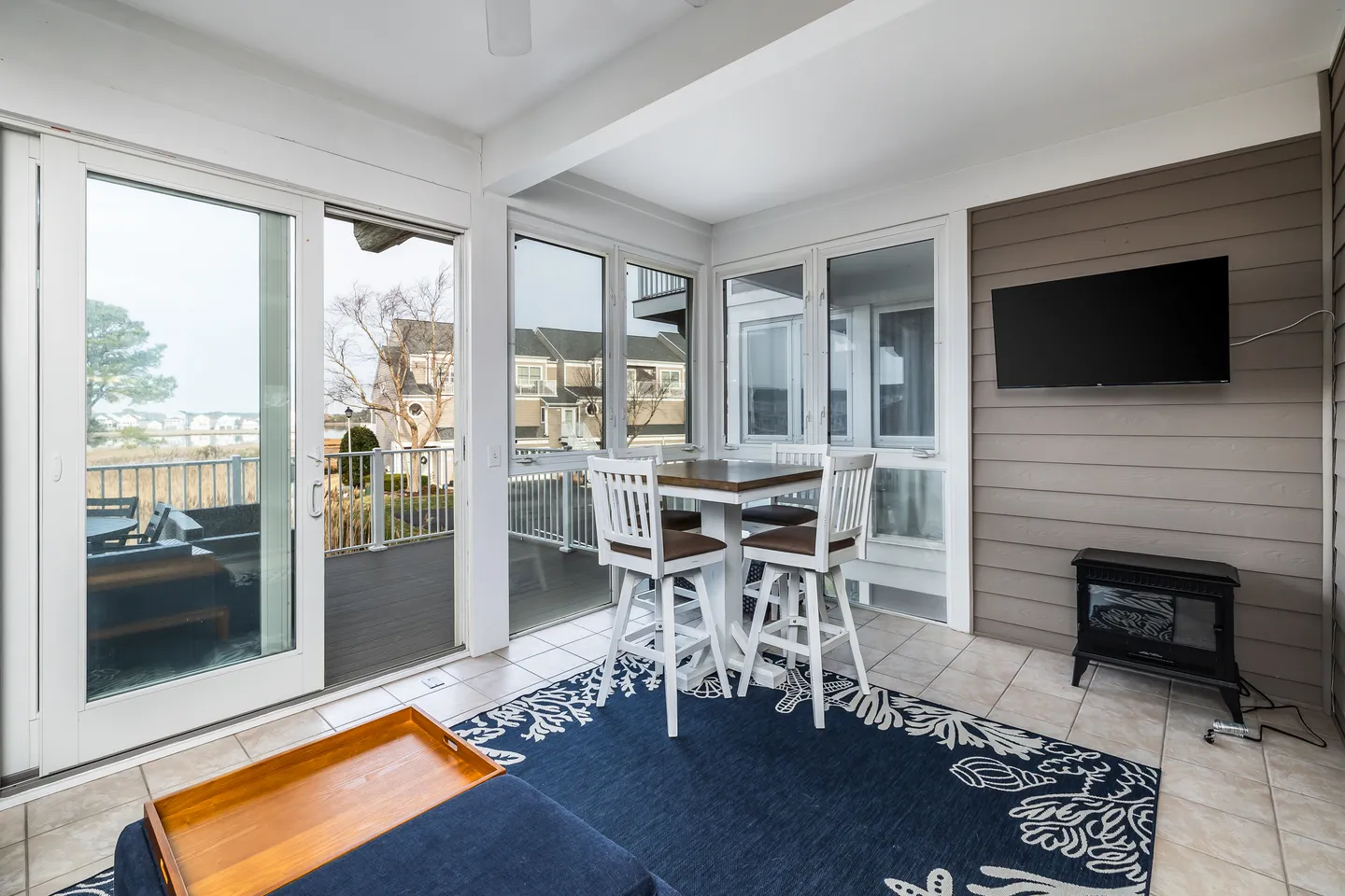 Sunroom with tiled floor, high-top table and barstools, wall-mounted television, and sliding glass doors opening to an outdoor deck.