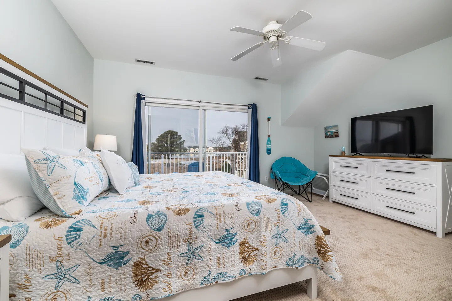 Bedroom with coastal-themed bedding, a ceiling fan, a large dresser with a TV, and sliding glass doors leading to a balcony.